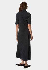 Schwarzes, kurzärmliges Kleid mit Stehkragen und fließender Silhouette bis zur Wade. Glatte Textur, minimalistisches Design, getragen mit dunkelblauen Ballerinas.