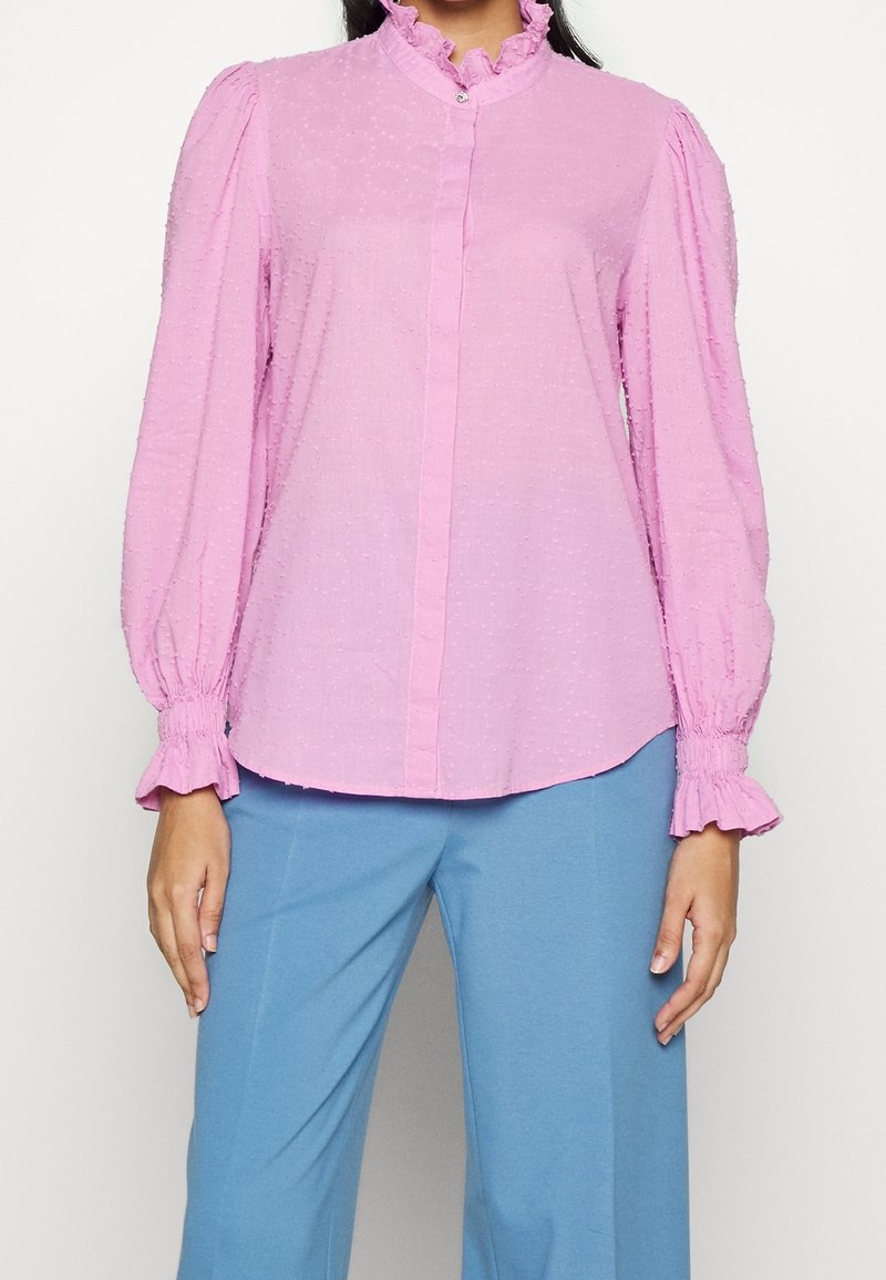 Femme portant un chemisier rose à manches longues avec col et poignets volantés, assorti à un pantalon bleu clair sur un fond neutre.