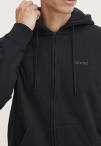 Svart hettegenser med glidelås laget av bomullsblanding. Har et logo på brystet, snor i hetta og frontlommer.