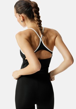 Frau mit geflochtenem Haar, die ein schwarzes Kleid mit weißen Trägern trägt, das einen Ausschnitt am oberen Rücken zeigt, posiert mit den Händen in der Hüfte.