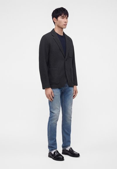 Homme portant un blazer sombre, une chemise noire, un jean bleu et des chaussures habillées noires, debout devant un fond clair uni.