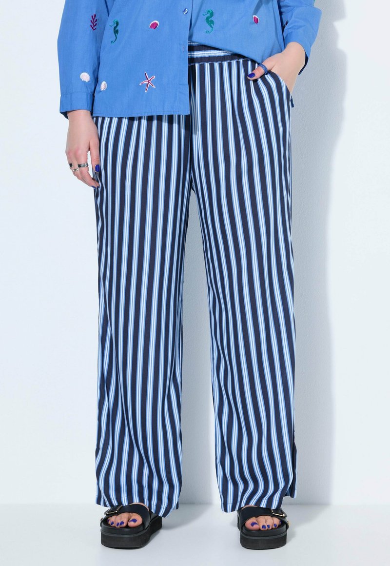 Person trägt lockere dunkelblaue Hose mit weißen vertikalen Streifen, blaues Hemd mit Stickerei von Meeresbewohnern und schwarze Sandalen mit Plateausohle.