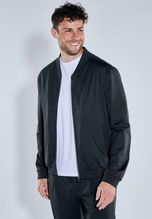 Black bomber jacket with a zip front, ribbed cuffs, and a smooth texture. Worn over a white crewneck t-shirt and paired with black pants.