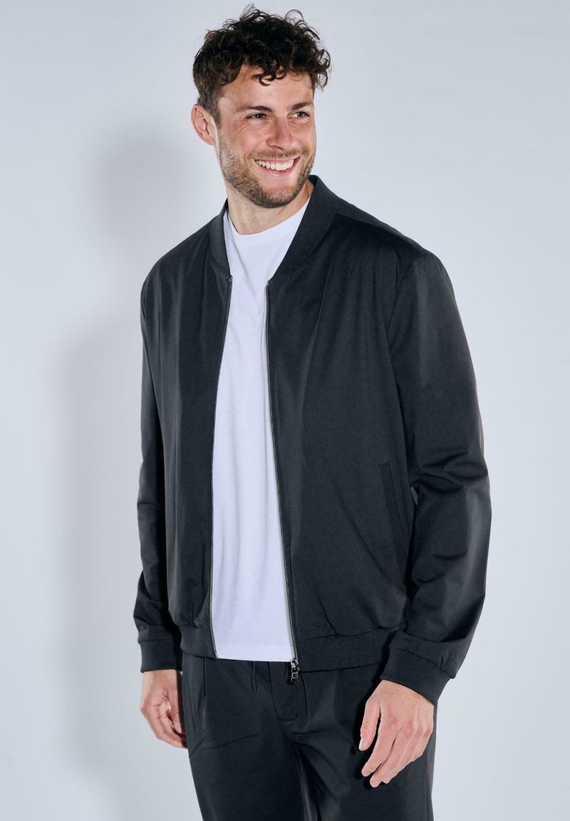 Schwarze Bomberjacke mit Reißverschluss, gerippten Bündchen und glatter Textur. Getragen über einem weißen Rundhals-T-Shirt und kombiniert mit schwarzen Hosen.