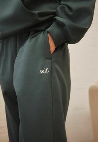 Donkergroene sweatpants van zacht materiaal met een zijzak en geborduurde tekst "self." op de dij. Elastische tailleband en manchetten.