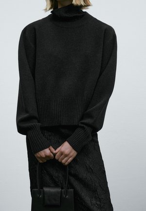 Person trägt einen schwarzen Pullover und Rock und hält eine kleine schwarze Handtasche mit beiden Händen vor einem einfarbigen Hintergrund.