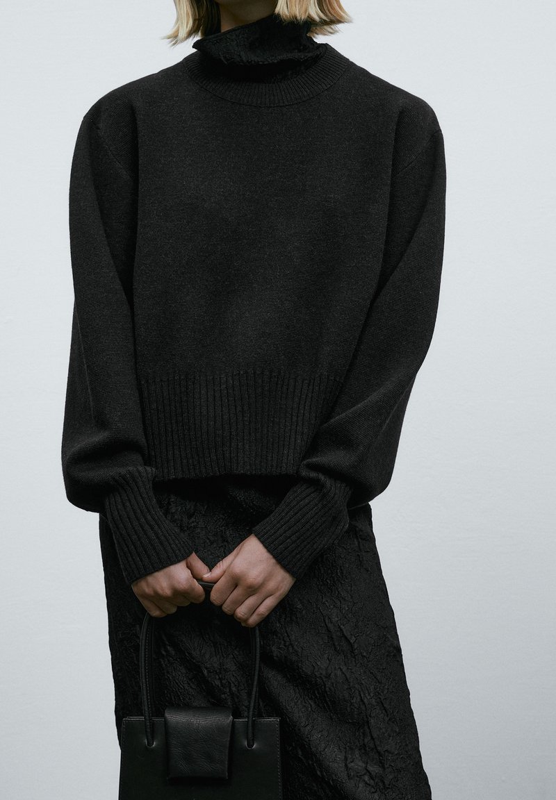 Personne portant un pull noir et une jupe, tenant un petit sac à main noir à deux mains sur un fond uni.