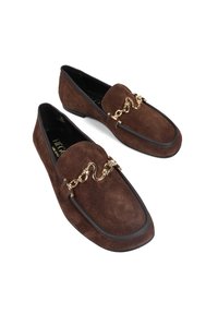 Brązowe zamszowe loafersy o eleganckim, zaokrąglonym kształcie, z detalem w postaci złotego łańcuszka na wierzchu stopy i czarnym wykończeniem wzdłuż podeszwy.