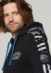 Sudadera con cremallera negra y cordones azules. Presenta texto y gráficos impresos en blanco en el pecho y la manga. Tela suave, ajuste estructurado.