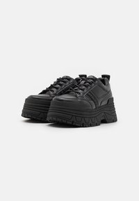 Zwarte platform sneaker met een chunky zool, uitgevoerd in glad leer, met ronde veters en reliëf logo accenten aan de zijkanten.