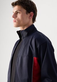 Ναυτικό μπουφάν με ψηλό γιακά, μπροστινό φερμουάρ και κόκκινη πλαϊνή ρίγα. Λείος ύφασμα με διακριτική γυαλάδα, featuring contrasting white stitching accents.