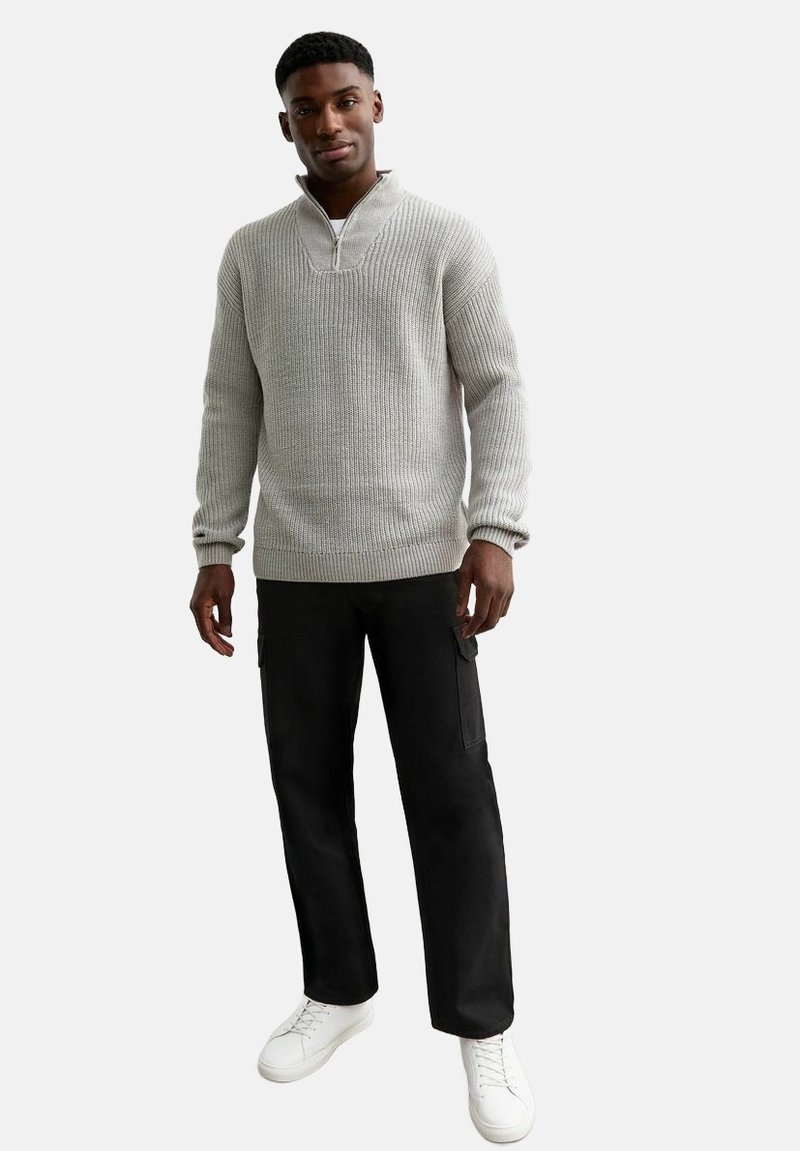 Suéter de punto acanalado en gris claro, con cuello de media cremallera, combinado con pantalones cargo negros y zapatillas blancas, creando un look informal.