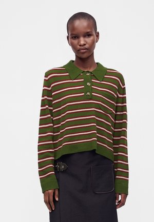 Femme aux cheveux courts porte un haut en maille à manches longues rayé vert, rouge et blanc avec des boutons, et une jupe noire avec une poche avant et un détail ceinture.