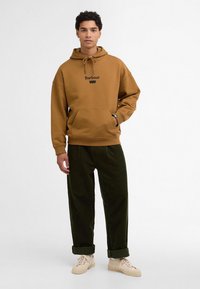 Καφέ φούτερ με μπροστινή τσέπη και μαύρο λογότυπο "Barbour", σε συνδυασμό με σκούρες πράσινες κοτλέ παντελόνες και μπεζ αθλητικά παπούτσια.