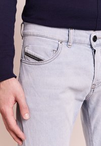 Muška ruka odpočívá poblíž kapsy světle modrých denimových jeansů s černou visačkou, na sobě má tmavý dlouhý rukáv.