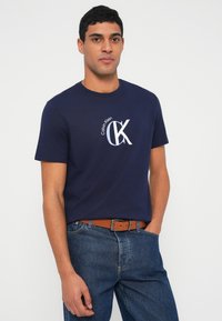 Tumši zila kokvilnas T-krekls ar apaļu kaklu, uz kura ir balts Calvin Klein logotips. Nēsāts kopā ar zilām džinsa biksēm un brūnu ādas jostu.