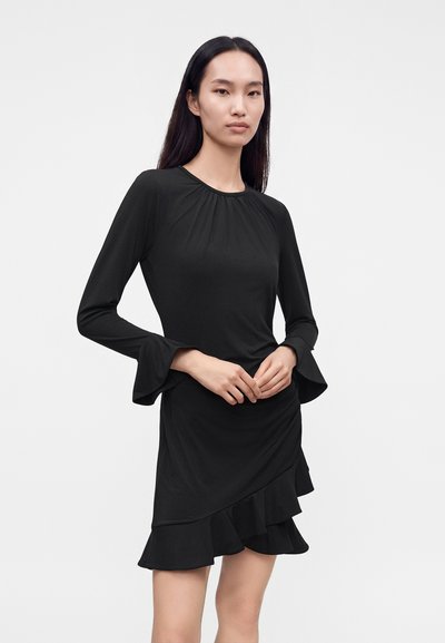 Femme aux longs cheveux foncés portant une robe noire à manches longues avec des poignets à volants et un ourlet volanté asymétrique, debout devant un fond uni.