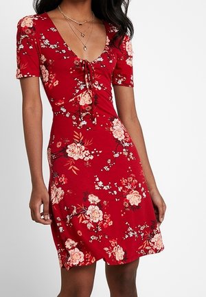 Femme portant une robe rouge fleurie à manches courtes, un décolleté en V avec un détail noué, et des colliers dorés superposés.