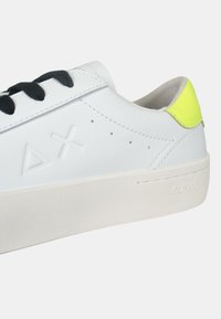 Sneaker bianco in pelle con lacci neri, superficie testurizzata, logo embossato e un accento giallo brillante sul tallone.