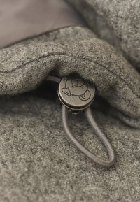 Hall villane kangas tekstuurse viimistlusega, millel on ümmargune metallnupp karu kujutisega ja hall nöör.