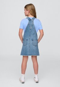 Fiatal személy áll háttal, világoskék inget, farmer kismama ruhát hátsó zsebekkel, fehér zoknit és fehér magas szárú edzőcipőt visel.