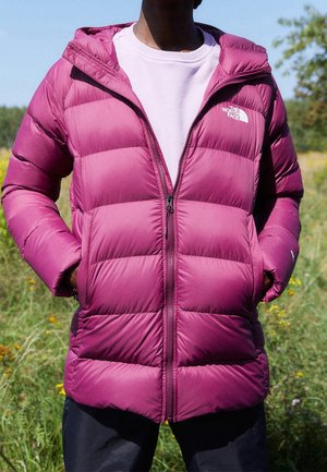 Personne portant une doudoune violette avec les mains dans les poches, debout à l'extérieur dans un champ d'herbe sous un ciel dégagé.