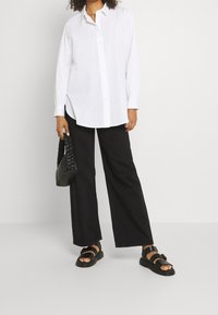 Camicia bianca con bottoni e maniche lunghe, abbinata a pantaloni neri a gamba larga e sandali neri. Include una piccola borsa intrecciata nera.