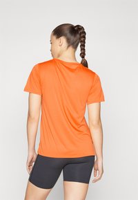 Oranžna športna majica s kratkimi rokavi in smooth teksturo, okroglim izrezom in sproščenim krojem, v kombinaciji s črnimi oprijetimi hlačami.
