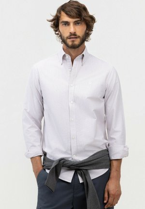Hombre con cabello rizado y castaño y barba, que lleva una camisa blanca de rayas con botones y un suéter gris atado alrededor de su cintura.