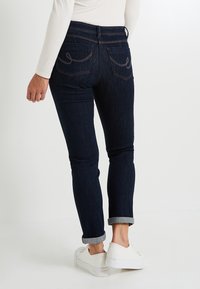 Jean en denim bleu foncé avec une coupe ajustée, ourlets retroussés et poches arrière courbées ornées de coutures décoratives. Associé avec des chaussures blanches.