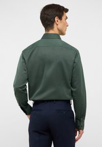 Donkergroen shirt van een gestructureerde stof, met knoopmanchetten en een standaardkraag, gecombineerd met marineblauwe broeken. Achteraanzicht weergegeven.
