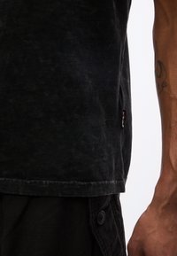 Gros plan sur une personne portant une chemise noire et un pantalon noir, avec un tatouage visible sur l'avant-bras.