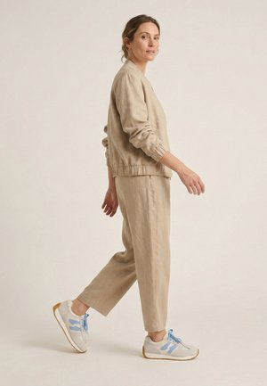 Vrouw loopt zijwaarts met beige linnen broek en bijpassend jasje met witte en blauwe sneakers tegen een effen beige achtergrond.