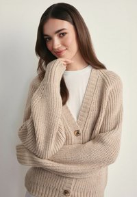 Beżowy sweter z dzianiny z głębokim dekoltem w kształcie V, oversize'owymi rękawami i dużymi guzikami. Teksturowana tkanina z prążkowanymi wzorami na całej powierzchni.