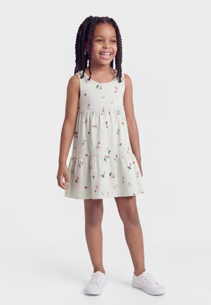 Χαμογελαστή νεαρή κοπέλα με πλεξούδες, που φοράει αμάνικο, ανοιχτόχρωμο φόρεμα με λουλουδένιο σχέδιο και λευκά αθλητικά παπούτσια, στέκεται σε λευκό φόντο.