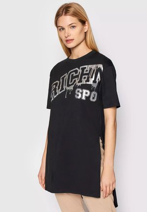 Donna con capelli biondi lisci che indossa una maglietta nera oversize con testo "RICH SPO" in argento che gocciola e pantaloni beige su uno sfondo semplice.