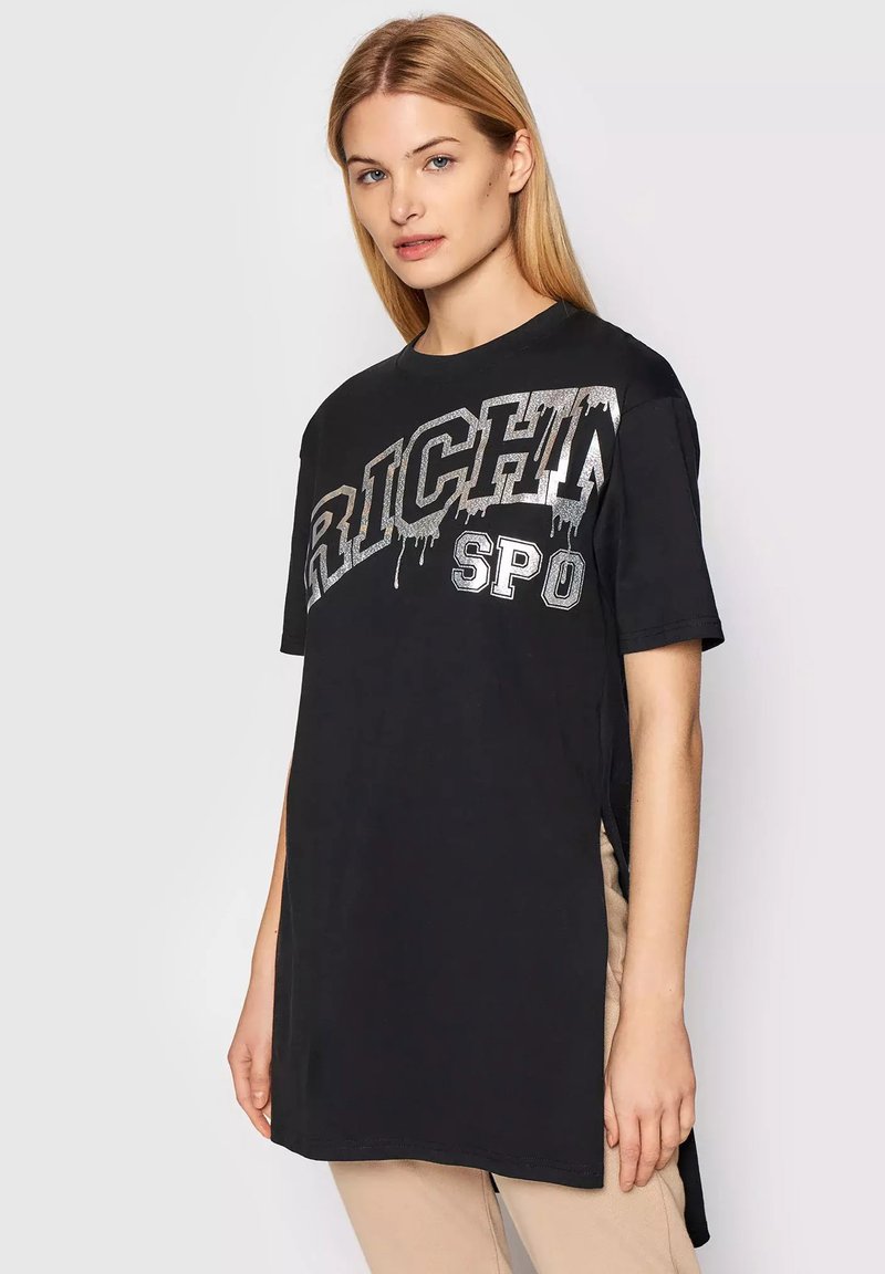 Donna con capelli biondi lisci che indossa una maglietta nera oversize con testo "RICH SPO" in argento che gocciola e pantaloni beige su uno sfondo semplice.