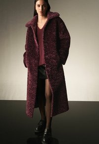 Overdimensioneret burgunder teddyfrakke med bred krave, forsynet med knapper, båret over en struktureret burgunder sweater og en sort mini nederdel. Sorte loafers.