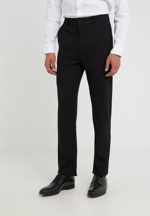 Mężczyzna ubrany w czarne, dopasowane spodnie garniturowe, czarne skórzane buty wizytowe oraz białą, długorękawową koszulę z guzikami, stojący na białej podłodze.