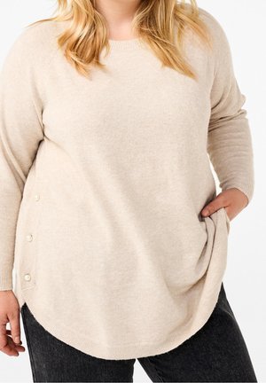 Kvinde iført en beige langærmet sweater med perleknapper på siden og sorte jeans, hånd i lommen.