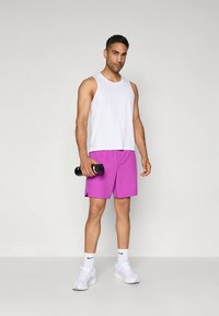 Top branco sem mangas, calções rosa-choque vibrantes, a segurar um haltere preto, ténis desportivos brancos, em pé sobre um fundo simples.