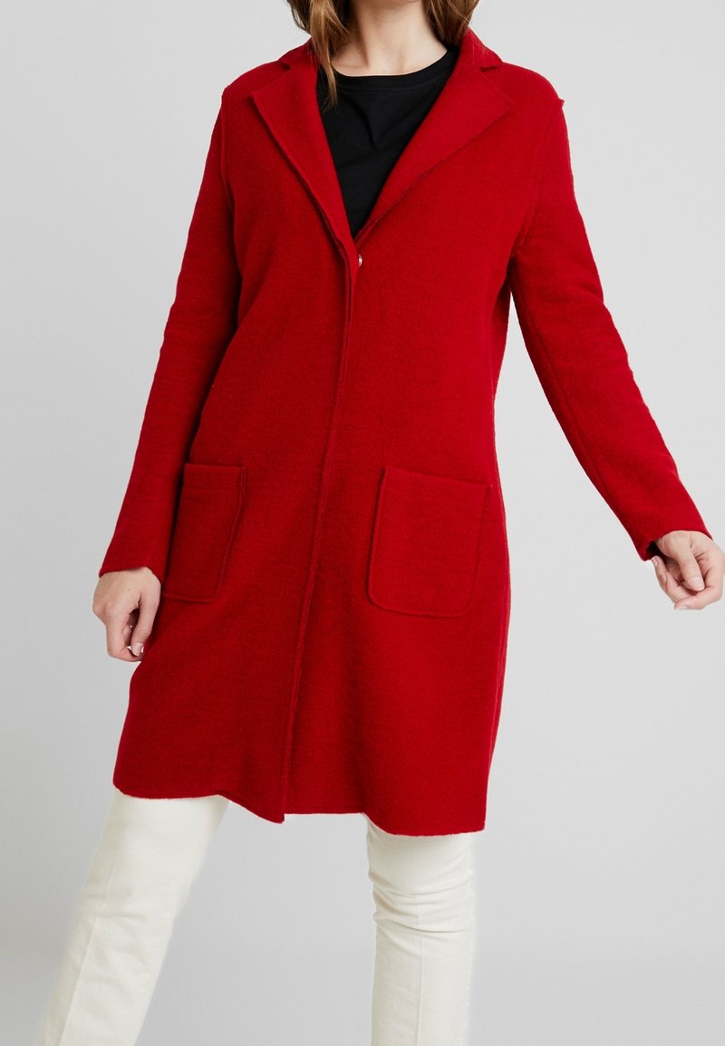 Röd ullkappa med krage, långa ärmar, två framfickor och rak skärning. Slät textur med en något borstad yta.