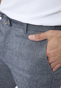 Pantalon en tissu tissé gris avec une finition texturée. Dispose d'une taille à cordon de serrage et de poches latérales. Main reposant sur la poche latérale.