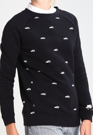 Czarna bluza z małymi białymi nadrukami tenisówek rozmieszczonymi na przodzie, noszona na białej koszuli z kołnierzykiem.