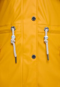 Jachetă galbenă impermeabilă cu accente argintii. Are șnururi împletite albe și gri cu capete din plastic. Două butoane circulare negre cu logo.