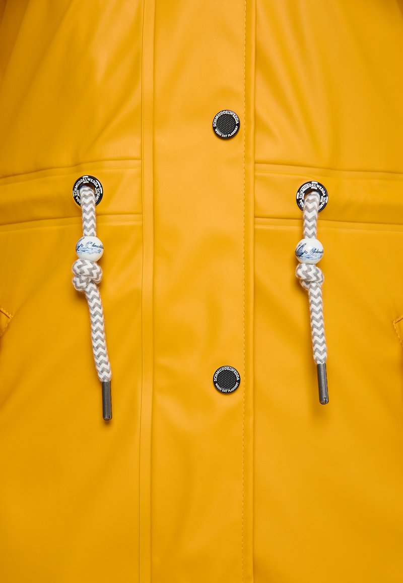 Geel waterdicht jack met zilveren accenten. Voorzien van witte en grijze gevlochten elastieken met plastic uiteinden. Twee ronde zwarte logoknoppen.