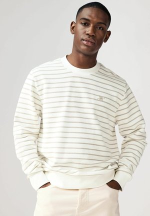 Sudadera blanca con franjas horizontales beiges, cuello redondo y puños acanalados. Presenta un pequeño logo bordado en el pecho.