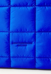Tissu matelassé bleu avec des surpiqûres carrées et "ARKK COPENHAGEN EST. 2015" brodé au centre.