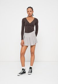 Woman with braided hair wearing a brown long-sleeve floral top, gray pleated mini skirt, white socks, and black Converse sneakers.