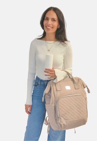 Mochila acolchada de color beige claro con asa doble, bolsillo frontal con cremallera y dos bolsillos laterales; mujer sosteniendo un recipiente de bebida aislante blanco.